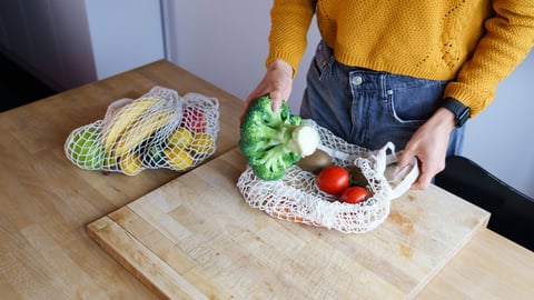 Woman holding broccoli and other produce in an eco-friendly mesh bag on a wooden table