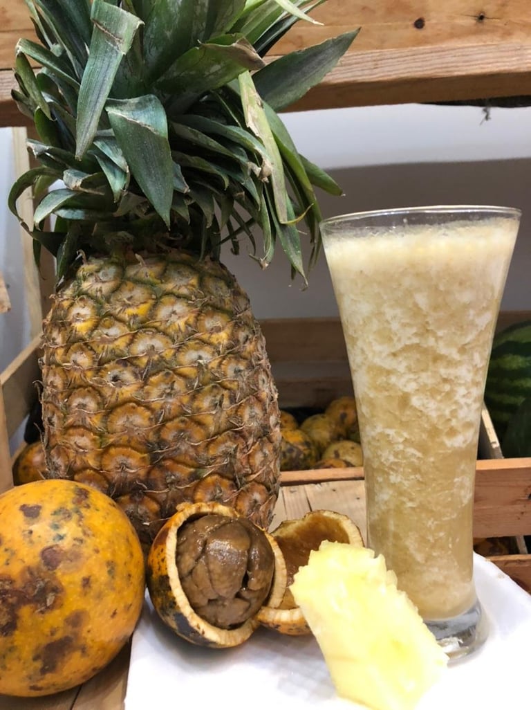 Fresh pineapple with leafy crown next to a tall glass of pineapple juice, surrounded by whole and cracked passion fruits on a wooden surface