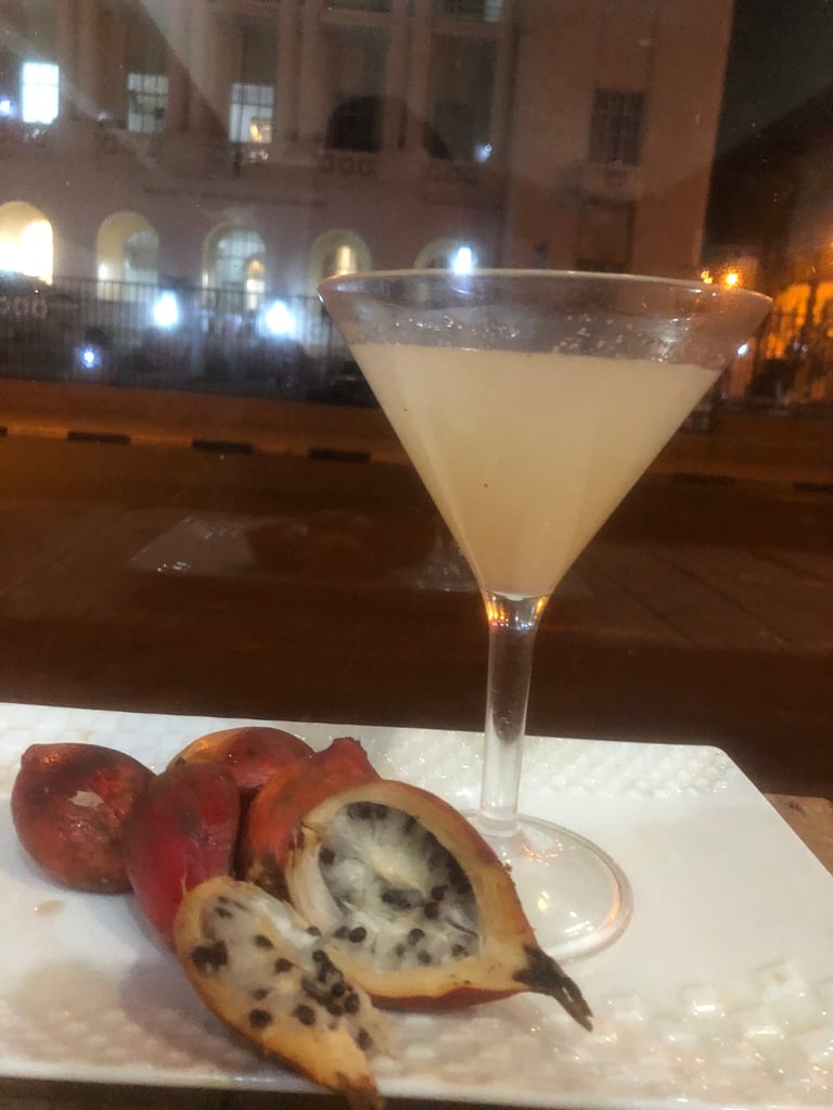 A pale cocktail in a martini glass beside sliced passion fruit on a white plate, photographed at night with illuminated buildings in background