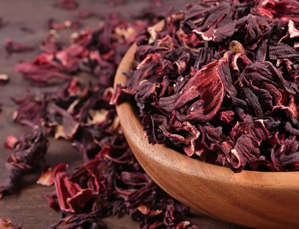 Dried hibiscus petals in a wooden bowl on a dark surface with scattered pieces